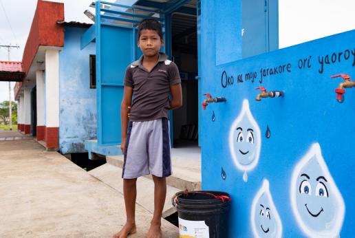 Un niño indígena parado al lado de un balde y una pared azul con gotas de agua pintadas.