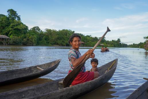 Mujer indígena junto a un niño pequeño en una canoa en el río.  Ambos miran a la cámara. De fondo hay más personas indígenas navegando en canoa.