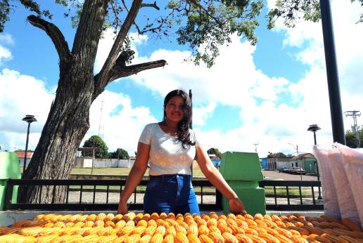 Una mujer sonríe a cámara parada detrás de una mesa con gran cantidad de cultivo de maíz.