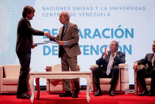 El Coordinador Residente de la ONU en Venezuela y el Rector de la Universidad Central de Venezuela se dan un apretón de manos en un escenario frente a una pantalla.