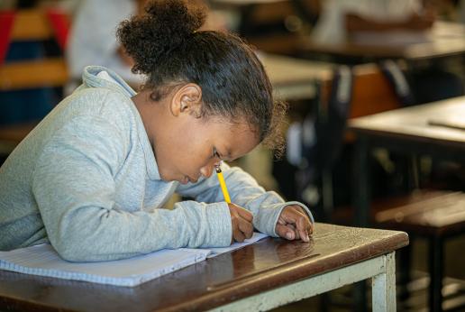 Niña escribe en un cuaderno sentada en un pupitre escolar.