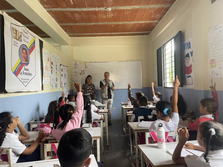 Un aula de clases con un grupo de niños alzando la mano y personal docente al fondo.