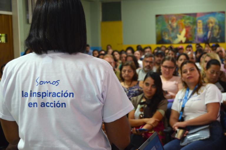 Una mujer con una franela del Programa UNV habla ante un público en un auditorio.