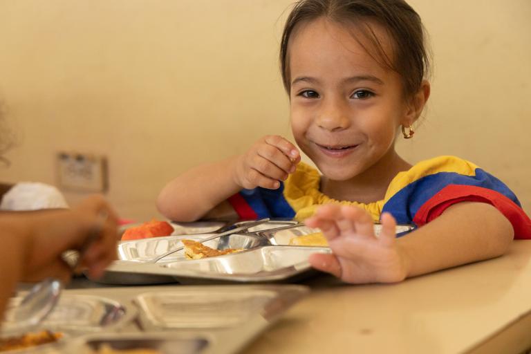 Niña sentada en una mesa come un plato de comida caliente.